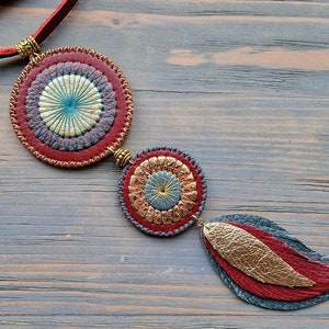 Puede incluir: Un collar de cuero con tres colgantes circulares. Los colgantes son rojos, azules y dorados con un dise&ntilde;o cosido. El collar est&aacute; colgado en una superficie de madera.