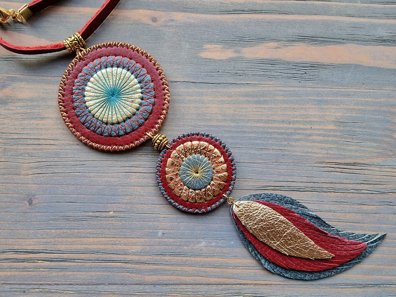 Puede incluir: Un collar de cuero con tres colgantes circulares. Los colgantes son rojos, azules y dorados con un dise&ntilde;o cosido. El collar est&aacute; colgado en una superficie de madera.