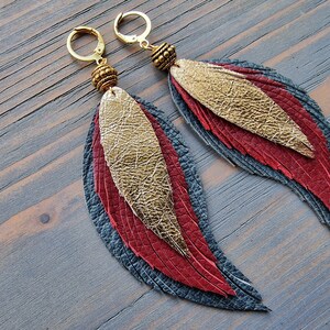 Puede incluir: Un par de aretes largos y colgantes con un dise&ntilde;o de plumas superpuestas. Las plumas est&aacute;n hechas de cuero en tonos grises, rojos y dorados. Los aretes tienen aros y cuentas doradas.