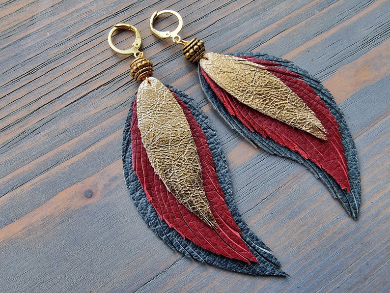 Puede incluir: Un par de aretes largos y colgantes con un dise&ntilde;o de plumas superpuestas. Las plumas est&aacute;n hechas de cuero en tonos grises, rojos y dorados. Los aretes tienen aros y cuentas doradas.
