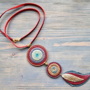 Puede incluir: Un collar de cuero rojo con tres colgantes circulares. Los colgantes est&aacute;n decorados con bordados dorados y azules. El colgante inferior tiene forma de pluma.