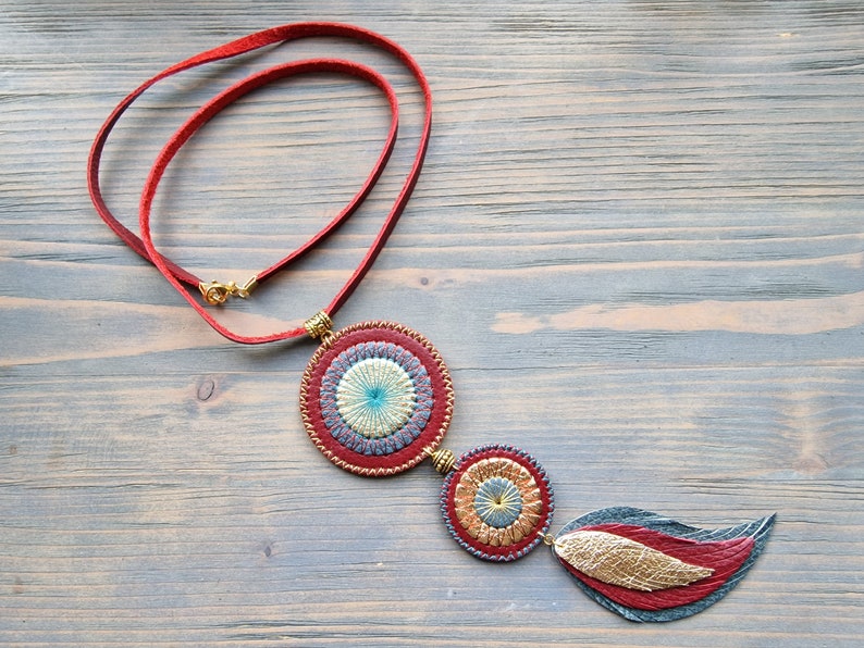 Puede incluir: Un collar de cuero rojo con tres colgantes circulares. Los colgantes est&aacute;n decorados con bordados dorados y azules. El colgante inferior tiene forma de pluma.