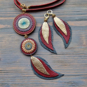Puede incluir: Un collar de cuero rojo con dos colgantes circulares y un dije de pluma. Los colgantes tienen un dise&ntilde;o dorado y azul. Tambi&eacute;n se muestran pendientes a juego con dijes de plumas.