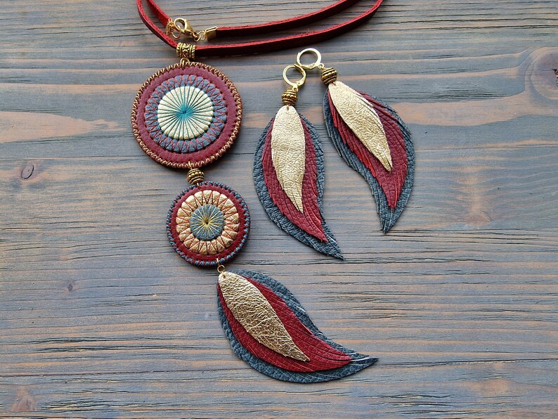 Puede incluir: Un collar de cuero rojo con dos colgantes circulares y un dije de pluma. Los colgantes tienen un dise&ntilde;o dorado y azul. Tambi&eacute;n se muestran pendientes a juego con dijes de plumas.