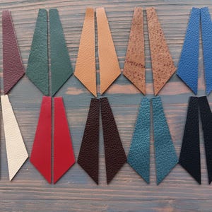 May include: A selection of leather earrings in shades of burgundy, green, tan, brown, blue, white, red, dark brown, teal, and black. Each pair features a geometric, elongated teardrop design, presented on a wooden surface.