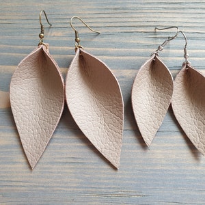 May include: Two pairs of leaf-shaped earrings in a light beige color. One pair has antique brass ear wires, and the other has silver-colored ear wires. The earrings have a textured surface and are displayed on a light blue wooden surface.
