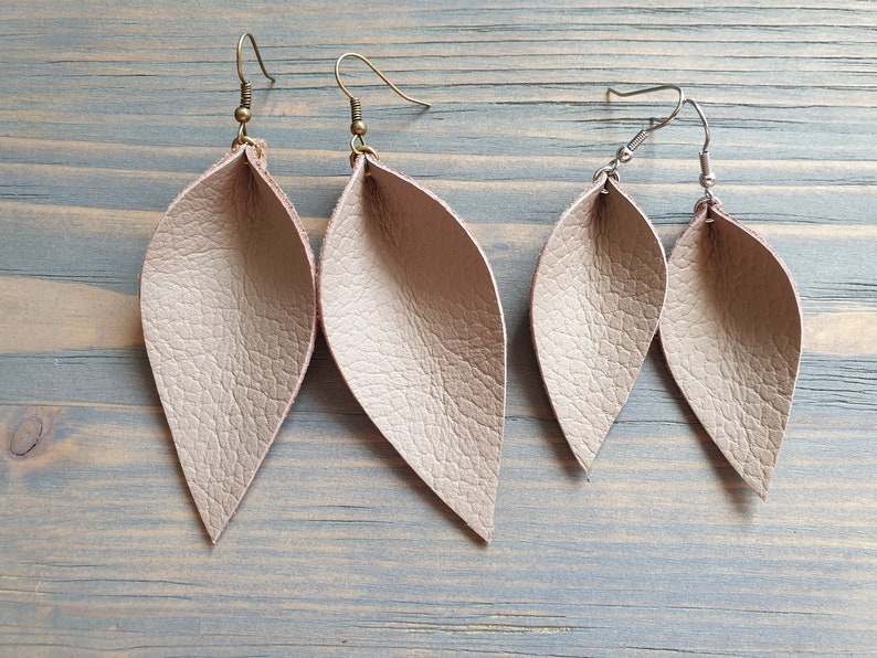May include: Two pairs of leaf-shaped earrings in a light beige color. One pair has antique brass ear wires, and the other has silver-colored ear wires. The earrings have a textured surface and are displayed on a light blue wooden surface.