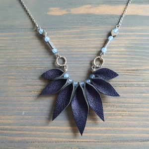 May include: A dark blue leather necklace with a geometric design. The necklace features a silver chain with blue beads and silver arrow-shaped charms. The leather pendant is made up of multiple overlapping leaves.