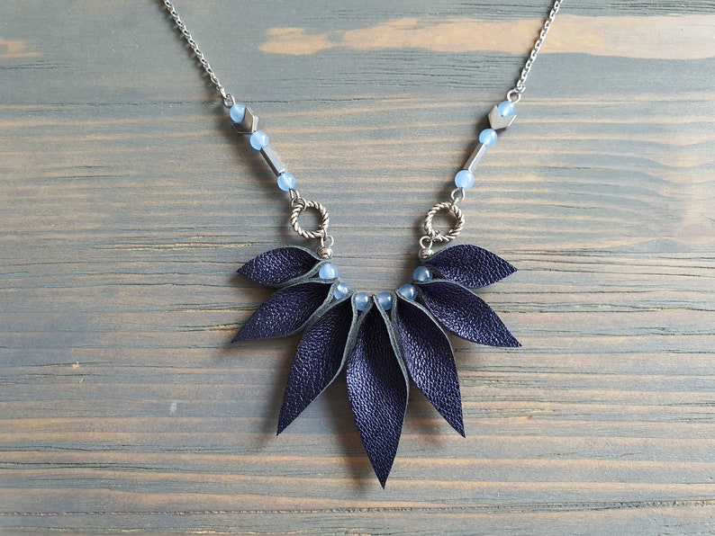 May include: A dark blue leather necklace with a geometric design. The necklace features a silver chain with blue beads and silver arrow-shaped charms. The leather pendant is made up of multiple overlapping leaves.