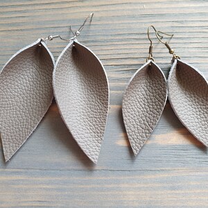 May include: Two pairs of gray leather leaf earrings. The earrings have a textured surface and are suspended from silver and bronze-colored ear wires. The earrings are displayed on a light blue wooden surface.