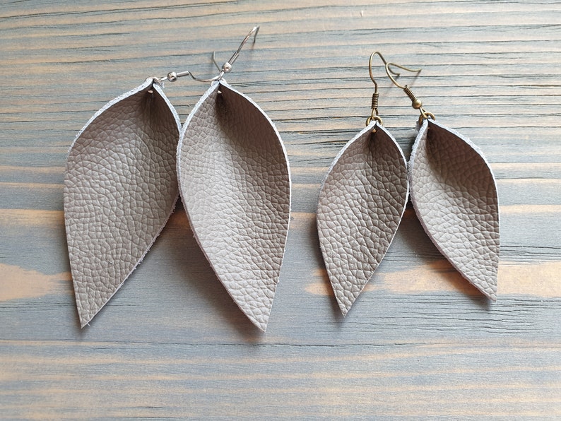 May include: Two pairs of gray leather leaf earrings. The earrings have a textured surface and are suspended from silver and bronze-colored ear wires. The earrings are displayed on a light blue wooden surface.