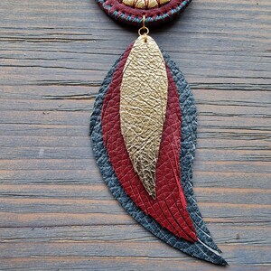 Puede incluir: Un pendiente de pluma de cuero en capas con un esquema de color dorado, rojo y azul oscuro. El pendiente cuelga de un bucle dorado.