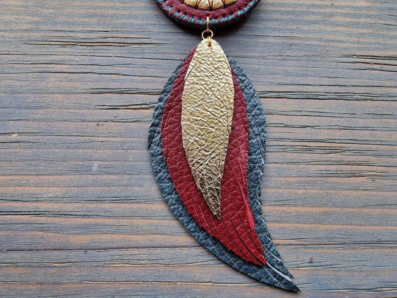 Puede incluir: Un pendiente de pluma de cuero en capas con un esquema de color dorado, rojo y azul oscuro. El pendiente cuelga de un bucle dorado.