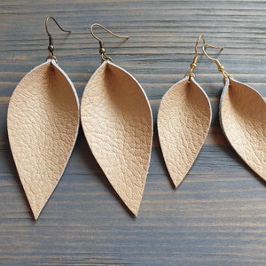 May include: Four pairs of leaf-shaped earrings in varying sizes. The earrings are a light gold color with a textured surface. The earring hooks are either bronze or gold-colored. The earrings are displayed on a weathered, gray-blue wooden surface.