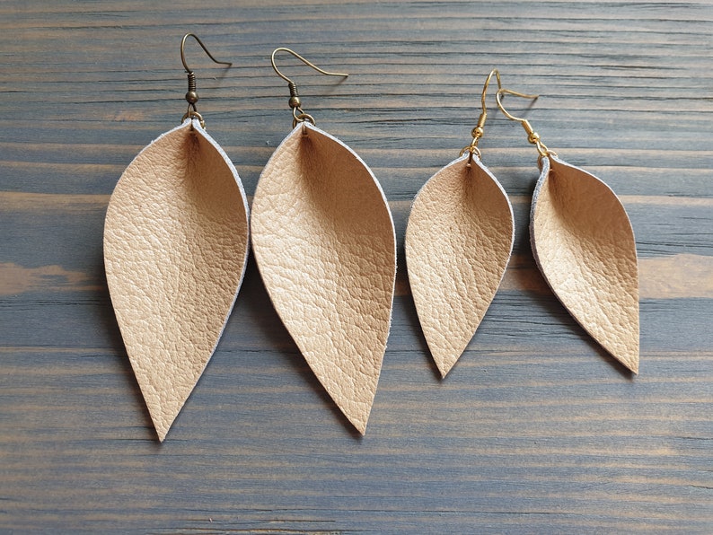 May include: Four pairs of leaf-shaped earrings in varying sizes. The earrings are a light gold color with a textured surface. The earring hooks are either bronze or gold-colored. The earrings are displayed on a weathered, gray-blue wooden surface.