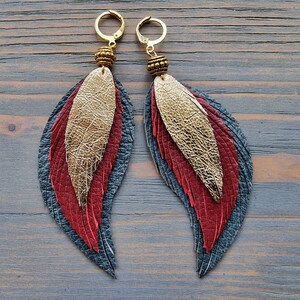 Puede incluir: Un par de pendientes colgantes con dise&ntilde;o de pluma. Los pendientes est&aacute;n hechos de cuero en tonos de rojo, dorado y gris. Los pendientes est&aacute;n adornados con cuentas doradas.