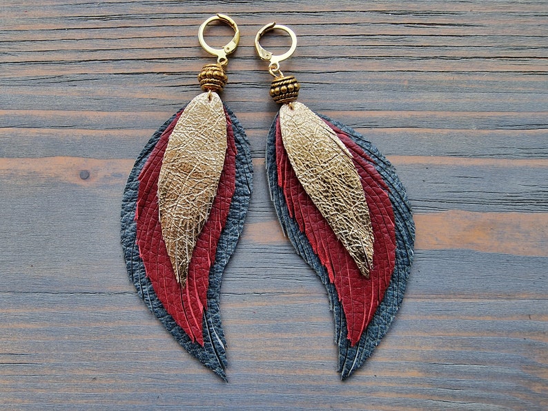 Puede incluir: Un par de pendientes colgantes con dise&ntilde;o de pluma. Los pendientes est&aacute;n hechos de cuero en tonos de rojo, dorado y gris. Los pendientes est&aacute;n adornados con cuentas doradas.