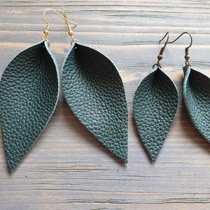 May include: Two pairs of leaf-shaped earrings. The larger pair has gold-colored ear wires, while the smaller pair has bronze-colored ear wires. Both pairs are made of textured, dark teal-colored leather. The earrings are displayed on a wooden surface.