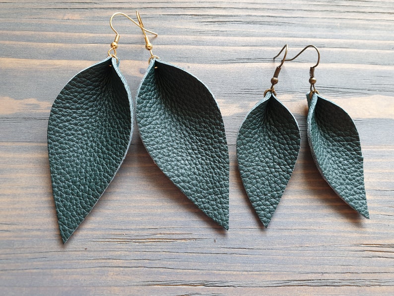 May include: Two pairs of leaf-shaped earrings. The larger pair has gold-colored ear wires, while the smaller pair has bronze-colored ear wires. Both pairs are made of textured, dark teal-colored leather. The earrings are displayed on a wooden surface.
