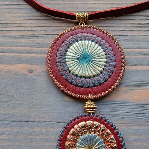 Puede incluir: Un collar de cuero rojo con dos colgantes circulares. Los colgantes est&aacute;n decorados con costuras doradas y azules, creando un patr&oacute;n de tipo mandala.
