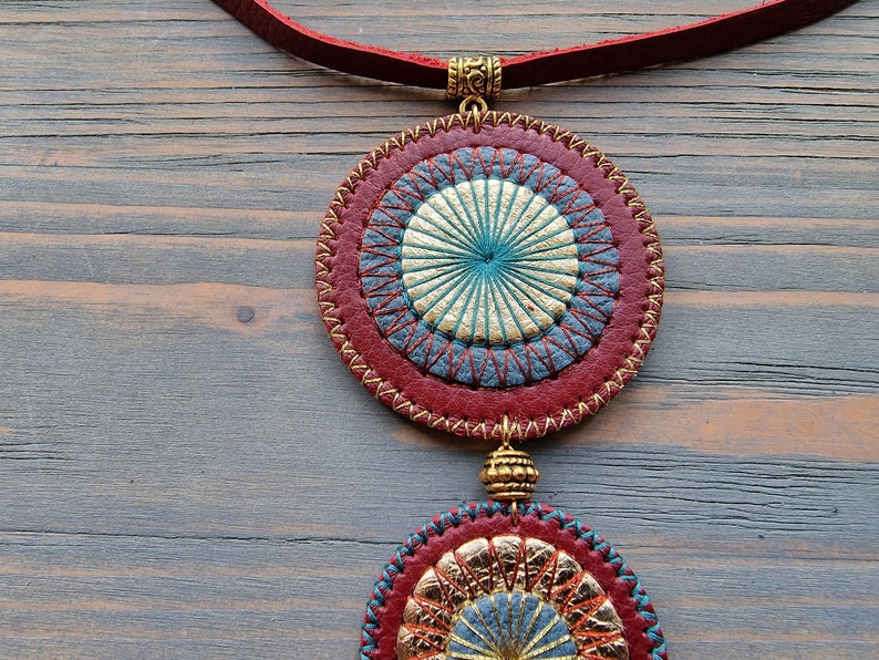 Puede incluir: Un collar de cuero rojo con dos colgantes circulares. Los colgantes est&aacute;n decorados con costuras doradas y azules, creando un patr&oacute;n de tipo mandala.