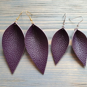 May include: Four pairs of purple leaf-shaped earrings. Two pairs have gold-colored hooks, and two pairs have silver-colored hooks. The earrings have a textured surface, and are arranged on a light blue wooden surface.