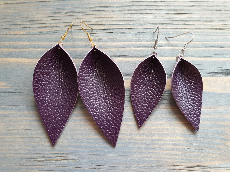 May include: Four pairs of purple leaf-shaped earrings. Two pairs have gold-colored hooks, and two pairs have silver-colored hooks. The earrings have a textured surface, and are arranged on a light blue wooden surface.