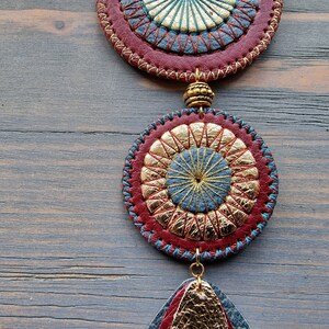 Puede incluir: Un colgante de cuero rojo, dorado y azul con un dise&ntilde;o circular y un detalle de flecos. El colgante est&aacute; colgado de una superficie de madera.