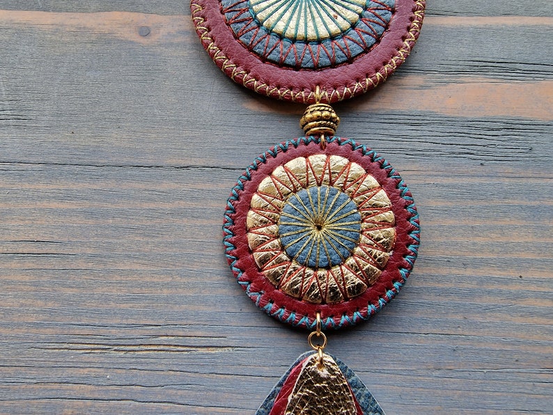 Puede incluir: Un colgante de cuero rojo, dorado y azul con un dise&ntilde;o circular y un detalle de flecos. El colgante est&aacute; colgado de una superficie de madera.
