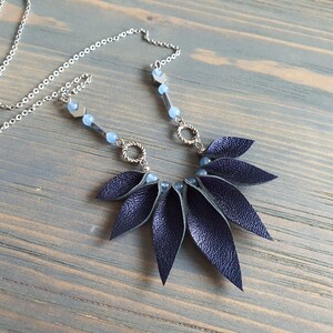 May include: A dark blue leather necklace with a silver chain and blue glass beads. The leather pendant is shaped like a fan with multiple overlapping leaves.