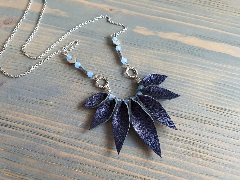 May include: A dark blue leather necklace with a silver chain and blue glass beads. The leather pendant is shaped like a fan with multiple overlapping leaves.