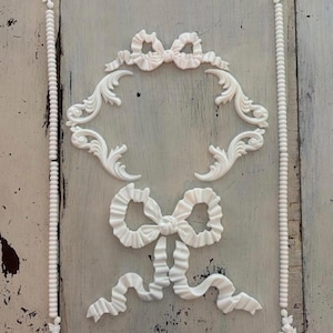 May include: White decorative wall art featuring a rectangular frame with floral corner accents and a beaded border. The design includes a bow at the top and a larger bow with flowing ribbons at the bottom, all in a classic, elegant style.