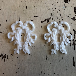 May include: Two white decorative keyhole covers with a floral and bow design. The covers are made of a smooth, white material and have a delicate, intricate design.