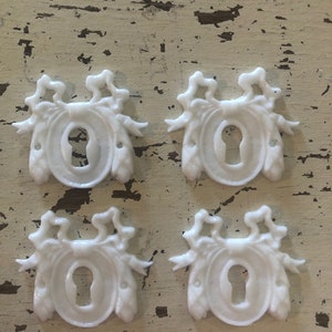 May include: Four white ceramic keyhole escutcheons with a decorative design. Each escutcheon features a stylized floral motif with a keyhole in the center.