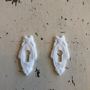 May include: Two white ceramic keyhole covers with ornate details. The keyhole covers are shaped like an oval with a keyhole in the center.