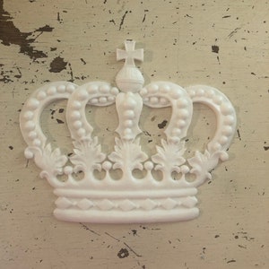 Puede incluir: Una corona decorativa blanca con una cruz en la parte superior. La corona tiene detalles ornamentados, incluyendo arcos con cuentas y detalles en forma de hojas. La corona está sobre un fondo blanco roto desgastado.