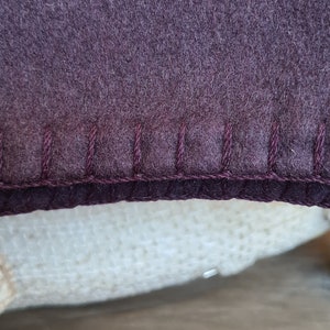 Op de afbeelding: Close-up van een donkerpaarse stof met een gestikte rand. De stof is zacht en pluche, en de stiksels zijn in een donkerdere tint paars.
