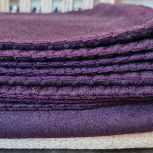 Op de afbeelding: Een stapel paarse fleece dekens met een witte stof eronder. De dekens zijn gevouwen en hebben een gestikte rand.