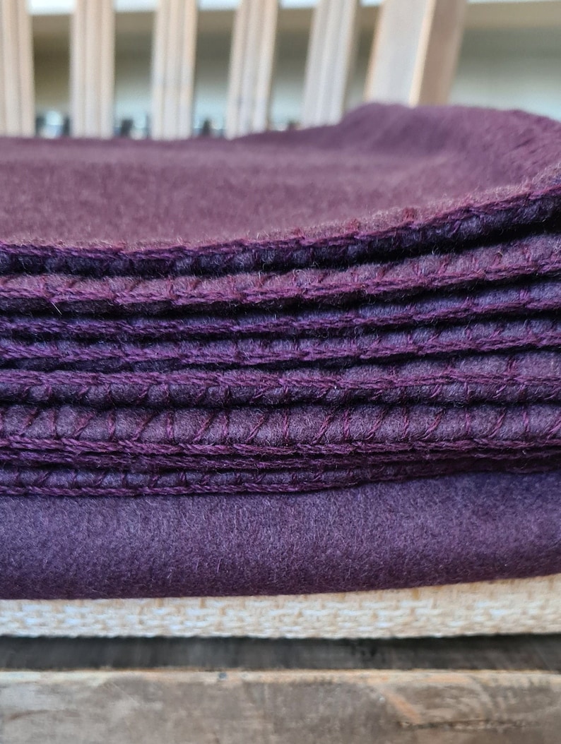Op de afbeelding: Een stapel paarse fleece dekens met een witte stof eronder. De dekens zijn gevouwen en hebben een gestikte rand.