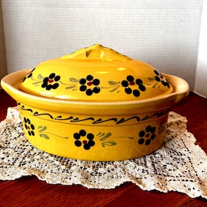 May include: A yellow ceramic casserole dish with a lid, decorated with black and red floral patterns and gray leaves. The dish has a handle on each side and sits on a white lace doily.