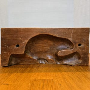 May include: A vintage wooden mold with a carved beaver design. The rectangular mold is made of aged, brown wood and features a detailed relief of a beaver sitting on a log. Two small holes are drilled into the wood.