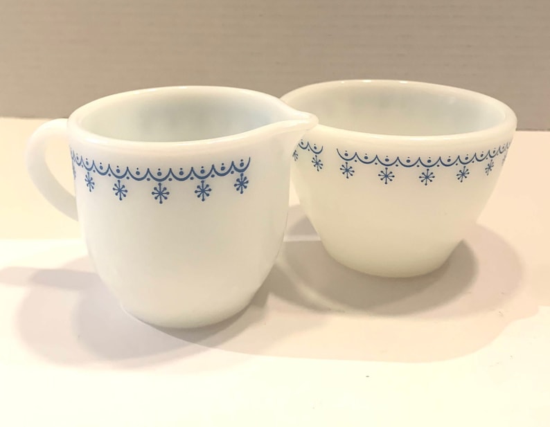 K&ouml;nnte beinhalten: Ein wei&szlig;es Milchk&auml;nnchen und eine Zuckerdose aus Milchglas mit einem blauen Schneeflockenmuster. Das Set hat einen Vintage-Look.