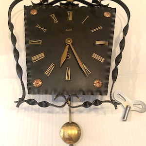 May include: A vintage black metal wall clock with gold Roman numerals and hands. The clock has a square face, a pendulum, and a winding key. Decorative elements include twisted metal and floral accents.