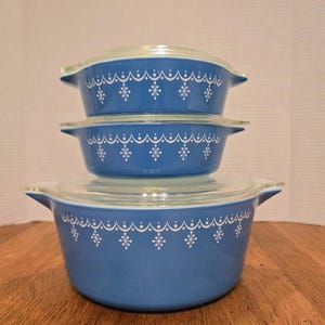May include: A stack of three blue Pyrex casserole dishes with clear glass lids. Each dish features a white snowflake and dotted design around the rim. The dishes are arranged on a wooden surface.