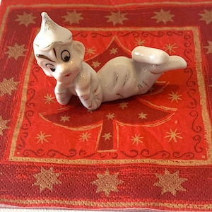 Peut inclure: Une figurine d'elfe en porcelaine blanche avec un chapeau pointu, allongée sur le côté sur une serviette à motif rouge et or. L'elfe a une expression espiègle et porte un chapeau blanc avec une bordure dorée.