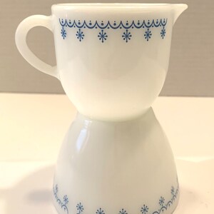 K&ouml;nnte beinhalten: Ein wei&szlig;er Milchglas-Sahnegie&szlig;er mit einem blauen Schneeflockenmuster. Der Sahnegie&szlig;er hat einen Griff und einen ausgestellten Boden.