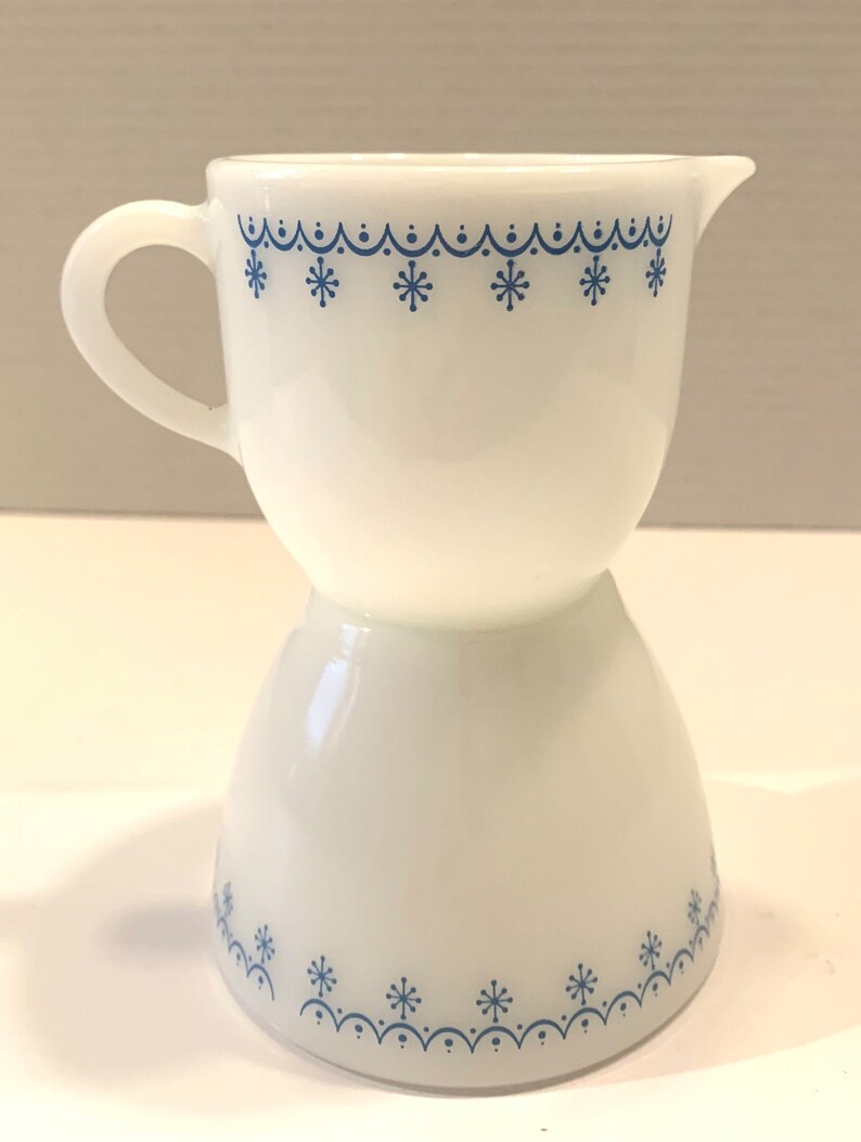 K&ouml;nnte beinhalten: Ein wei&szlig;er Milchglas-Sahnegie&szlig;er mit einem blauen Schneeflockenmuster. Der Sahnegie&szlig;er hat einen Griff und einen ausgestellten Boden.