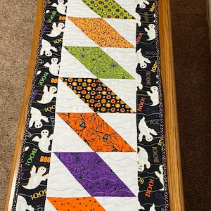 May include: A Halloween-themed table runner with a patchwork design. It features diagonal blocks in various patterns, including spiderwebs, polka dots, and Halloween motifs, set against a white background. The border is black with white ghosts and the word "BOO!"