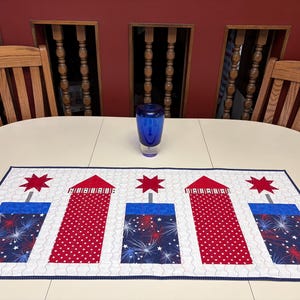 May include: A patriotic table runner with a white background, featuring red and blue firework designs. The runner has a navy blue border and is placed on a white oval table. A blue vase sits on the table. Wooden chairs and a red wall are in the background.