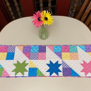 May include: A colorful quilted table runner with star and geometric patterns. The runner features squares of pink, purple, blue, and yellow fabric, with star shapes in green, blue, and pink. A vase with pink and yellow flowers sits on the table.